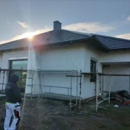 Dom jednorodzinny w trakcie ocieplania, widoczne rusztowania, pracownik w roboczym ubraniu i worki z materiałem izolacyjnym, jasne niebo z promieniami słonecznymi w tle.