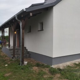 Len-Bud - Elewacja domu jednorodzinnego z drewnianą konstrukcją zadaszenia nad wejściem, świeżo pomalowana na biało z szarym cokołem, widoczne schody wejściowe z szarych płytek i wiadro po farbie obok jednej...