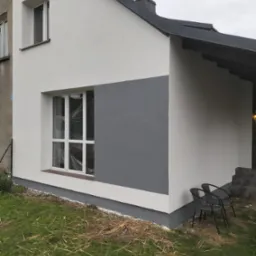 Fragment elewacji domu po remoncie, z białym i szarym tynkiem, okno zabezpieczone folią, dwa krzesła ogrodowe na trawniku.