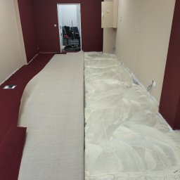 Len-Bud - Przygotowanie podłoża pod nową podłogę w biurze: szpachlowanie i układanie podkładu pod wykładzinę dywanową. Widoczne fragmenty bordowej ściany i beżowej ściany z puszką elektryczną.