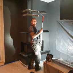 Len-Bud - Mężczyzna w kombinezonie malarskim trzyma wałek na teleskopowym kiju, malując ciemnoszarą ścianę w pokoju z osłoniętymi meblami i zamontowanymi półkami.