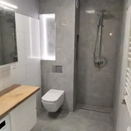 Nowoczesna łazienka wykończona w szarościach i bieli, z drewnianym blatem umywalki, podwieszaną toaletą i kabiną prysznicową typu walk-in z odpływem liniowym.