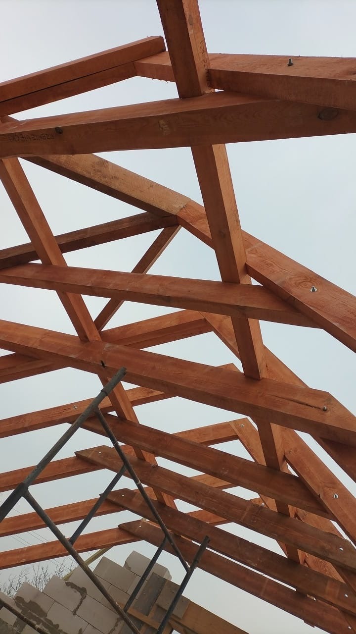 Drewniana konstrukcja dachu poddasza z widocznymi belkami i elementami mocującymi, częściowo zasłonięta rusztowaniem i blokami budowlanymi.