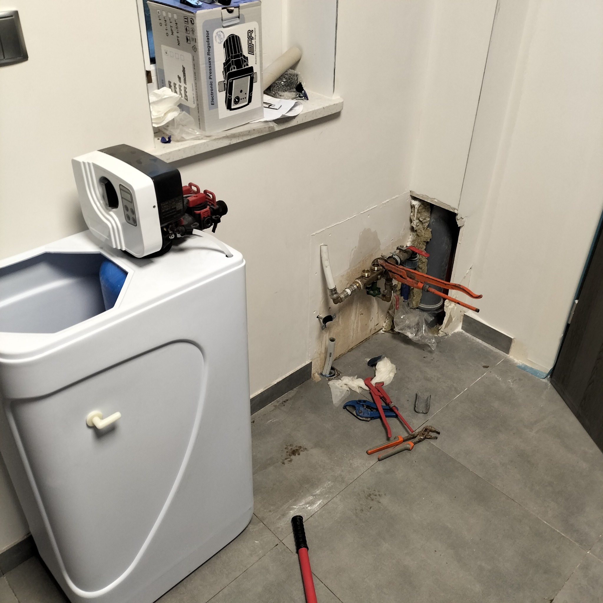Montaż zmiękczacza wody w łazience, widoczne podłączenia hydrauliczne w ścianie, narzędzia na podłodze i kartonowe opakowanie regulatora ciśnienia na parapecie okna.