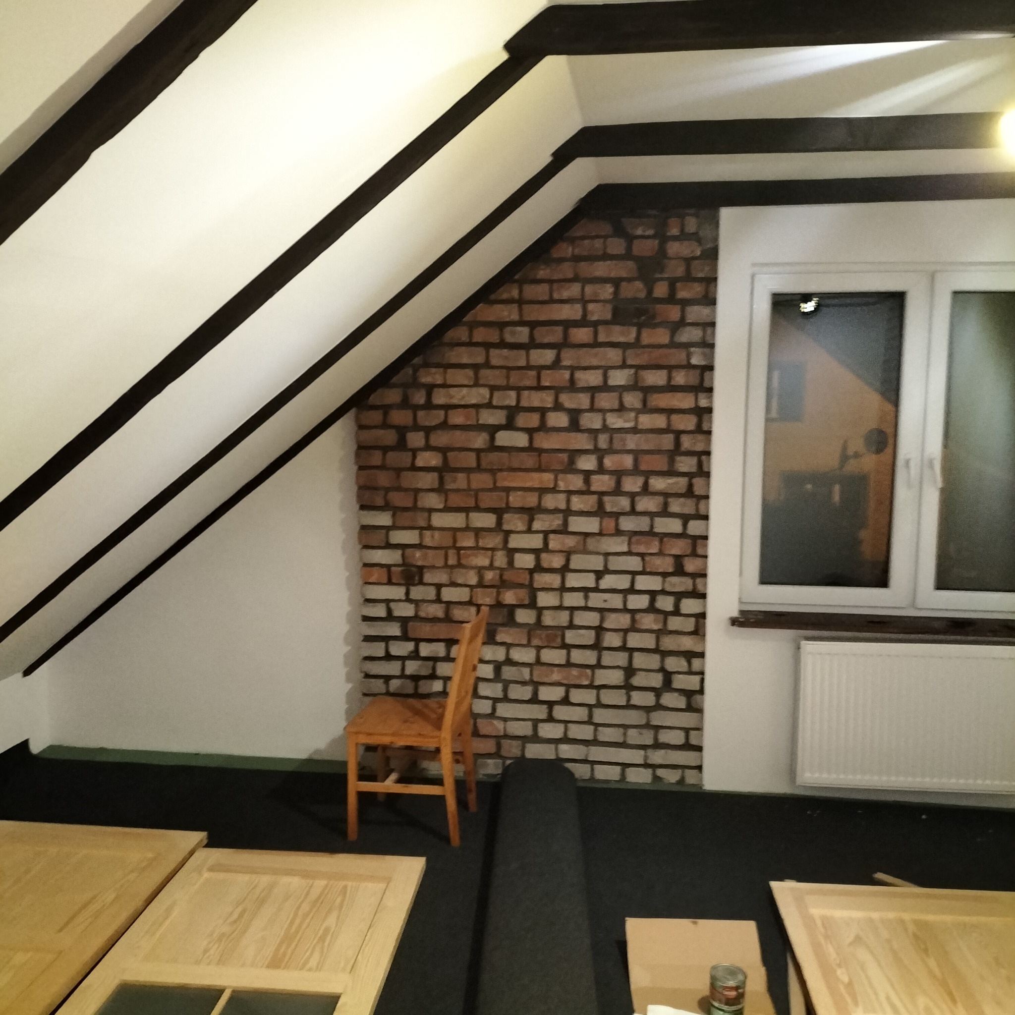 Poddasze z drewnianymi belkami, fragmentem ściany z cegły, oknem z grzejnikiem i drewnianym krzesłem. Na podłodze leżą drewniane elementy drzwi. Widoczna puszka farby.