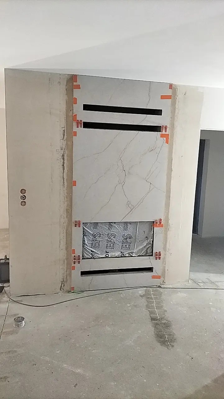 Ściana z marmurowymi płytami w trakcie montażu, z widocznymi otworami wentylacyjnymi i elementami mocującymi, zabezpieczona folią EPS w dolnej części, na surowym betonowym podłożu.