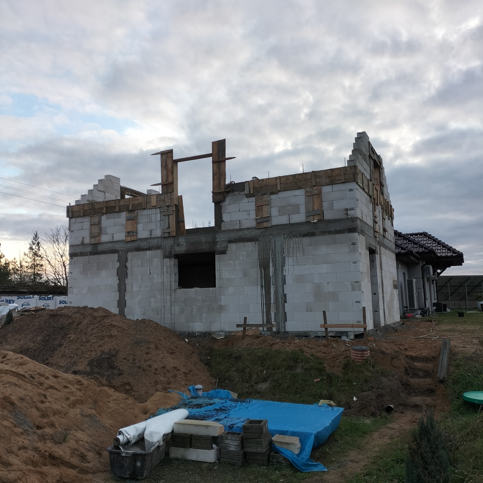 Budowa domu jednorodzinnego w trakcie wznoszenia ścian z bloczków betonowych, widoczne szalunki i stropy, w tle gotowy budynek z ciemnym dachem.