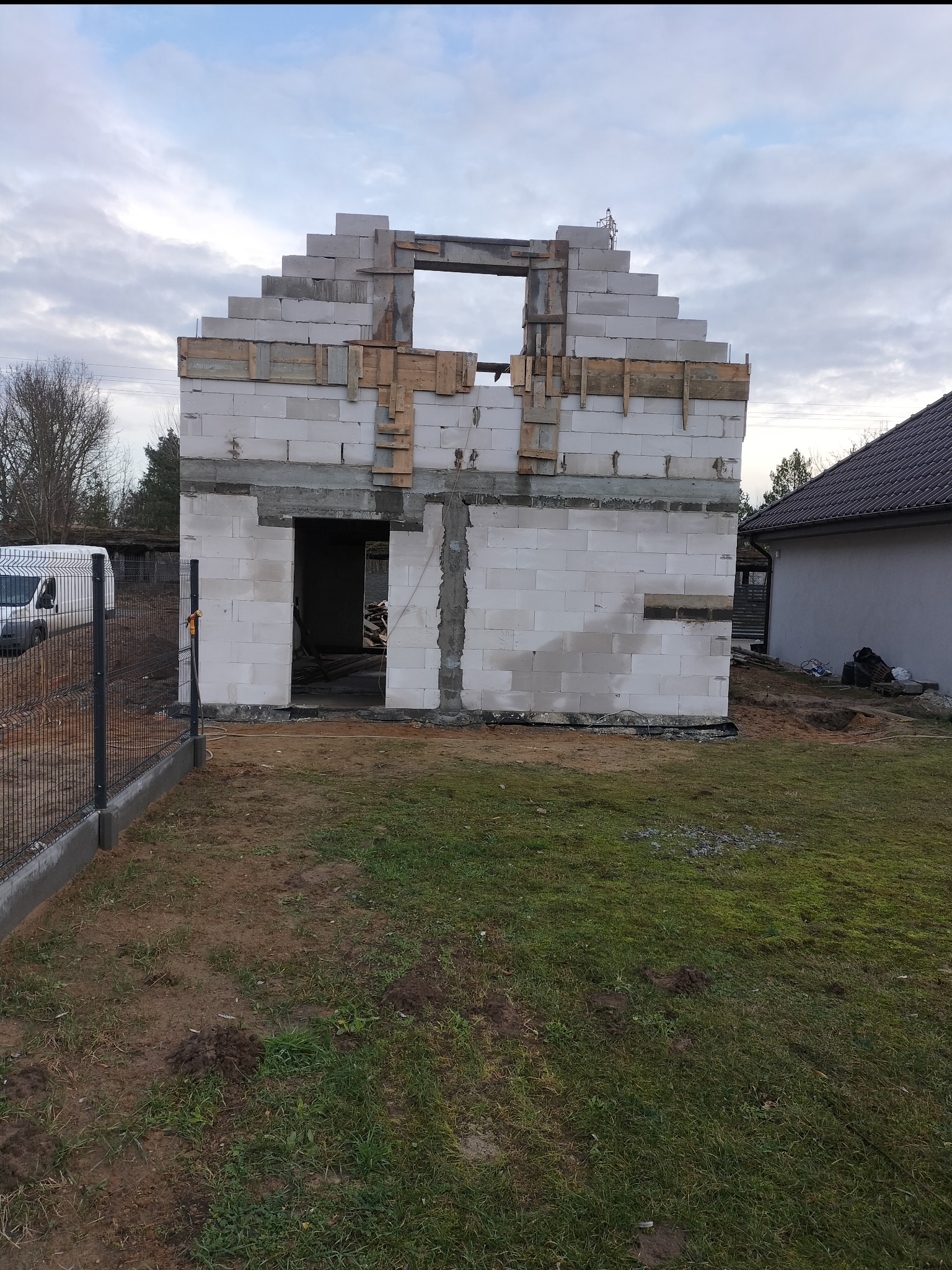 Surowy stan budynku w trakcie budowy z białych bloczków, widoczne drewniane szalunki wokół otworów okiennych i drzwiowych, częściowo ogrodzony teren budowy z widocznym samochodem dostawczym w tle.