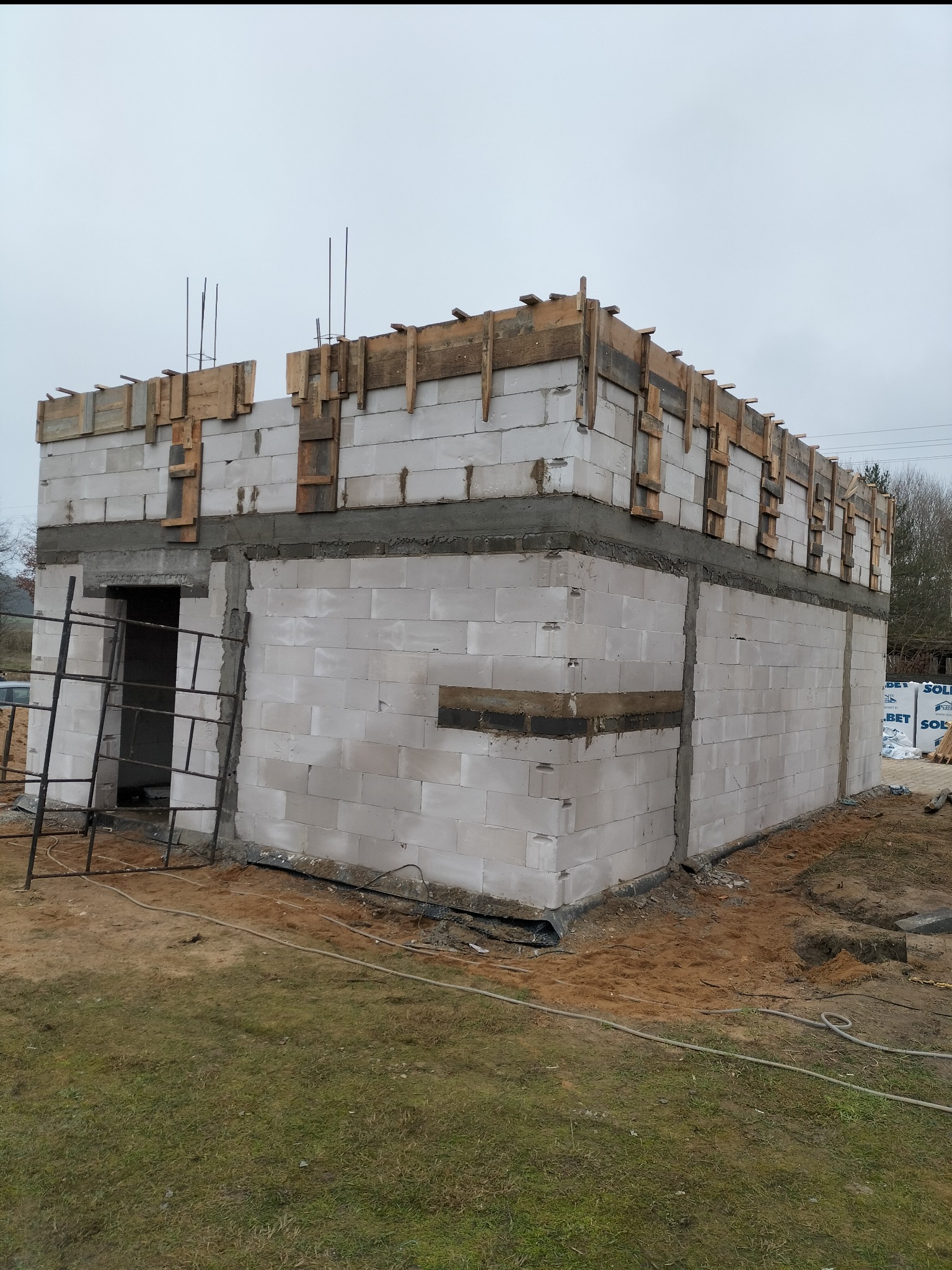 Surowy stan budynku z bloczków betonowych w trakcie budowy, z widocznym szalunkiem na stropie i wystającymi prętami zbrojeniowymi, na tle pochmurnego nieba.