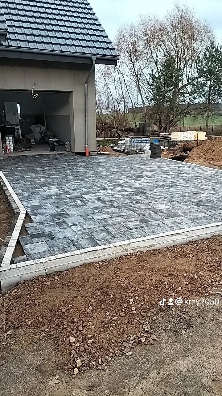 Układanie szarej kostki brukowej na podjeździe przed garażem, widoczne palety z kostką i przygotowane podłoże.