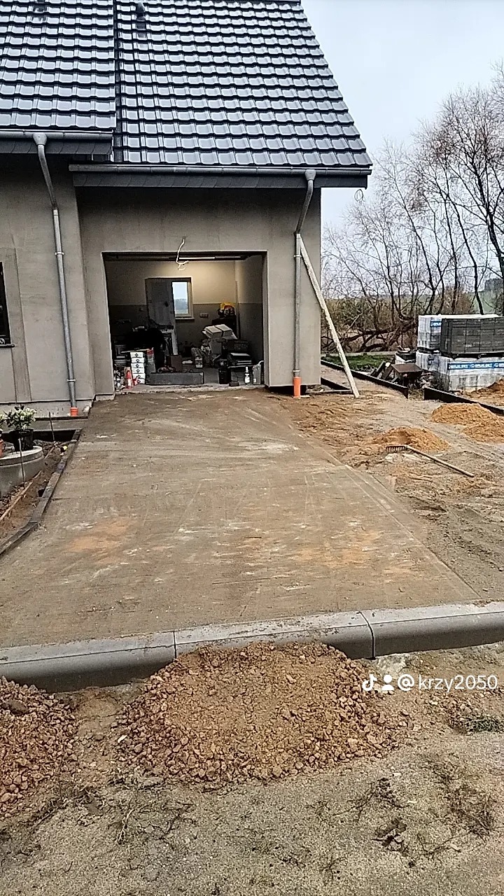 Betonowa podjazd do garażu w trakcie budowy, widoczne krawężniki, piasek i narzędzia budowlane, w tle garaż z materiałami budowlanymi