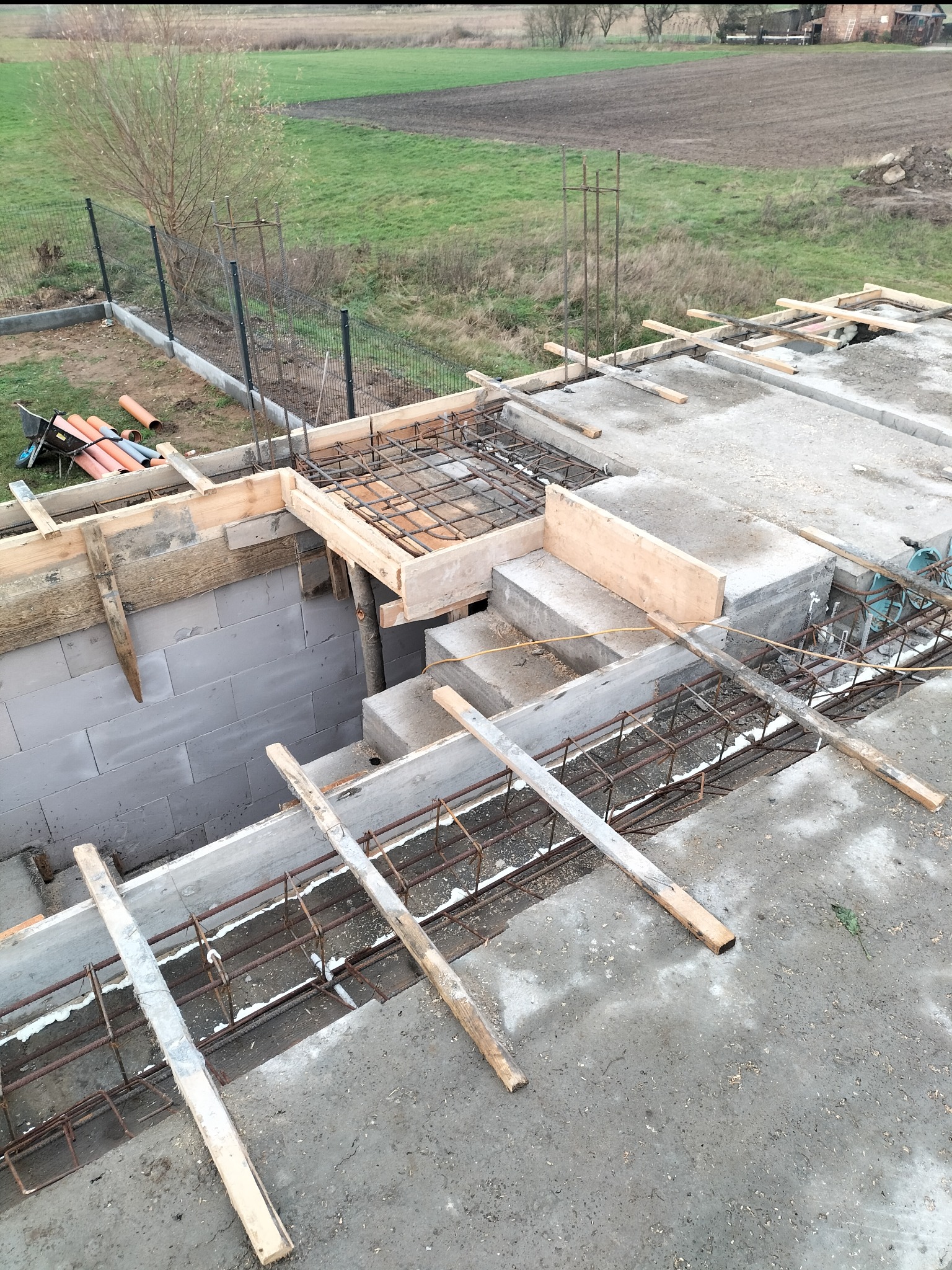 Konstrukcja z betonu i zbrojenia w trakcie budowy, widoczne drewniane szalunki i elementy stalowe, w tle ogrodzenie siatkowe i pole uprawne.