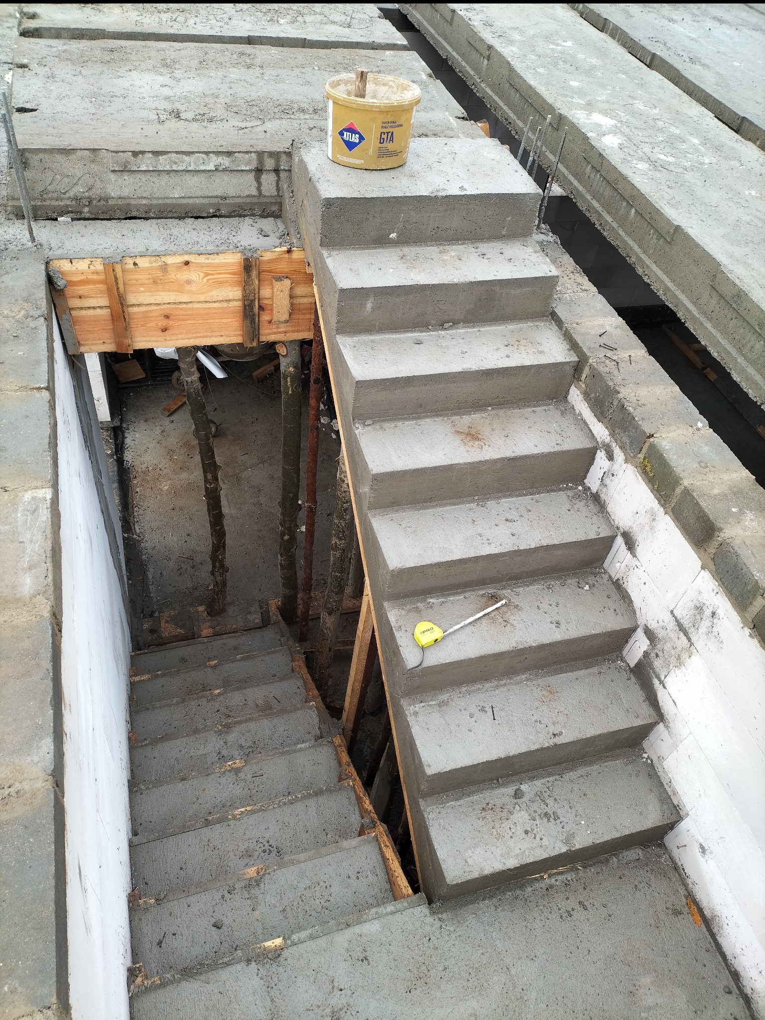 Surowe betonowe schody w trakcie budowy w Smogórach, widoczne drewniane szalunki i wiadro z zaprawą, żółta miara leży na jednym ze stopni.