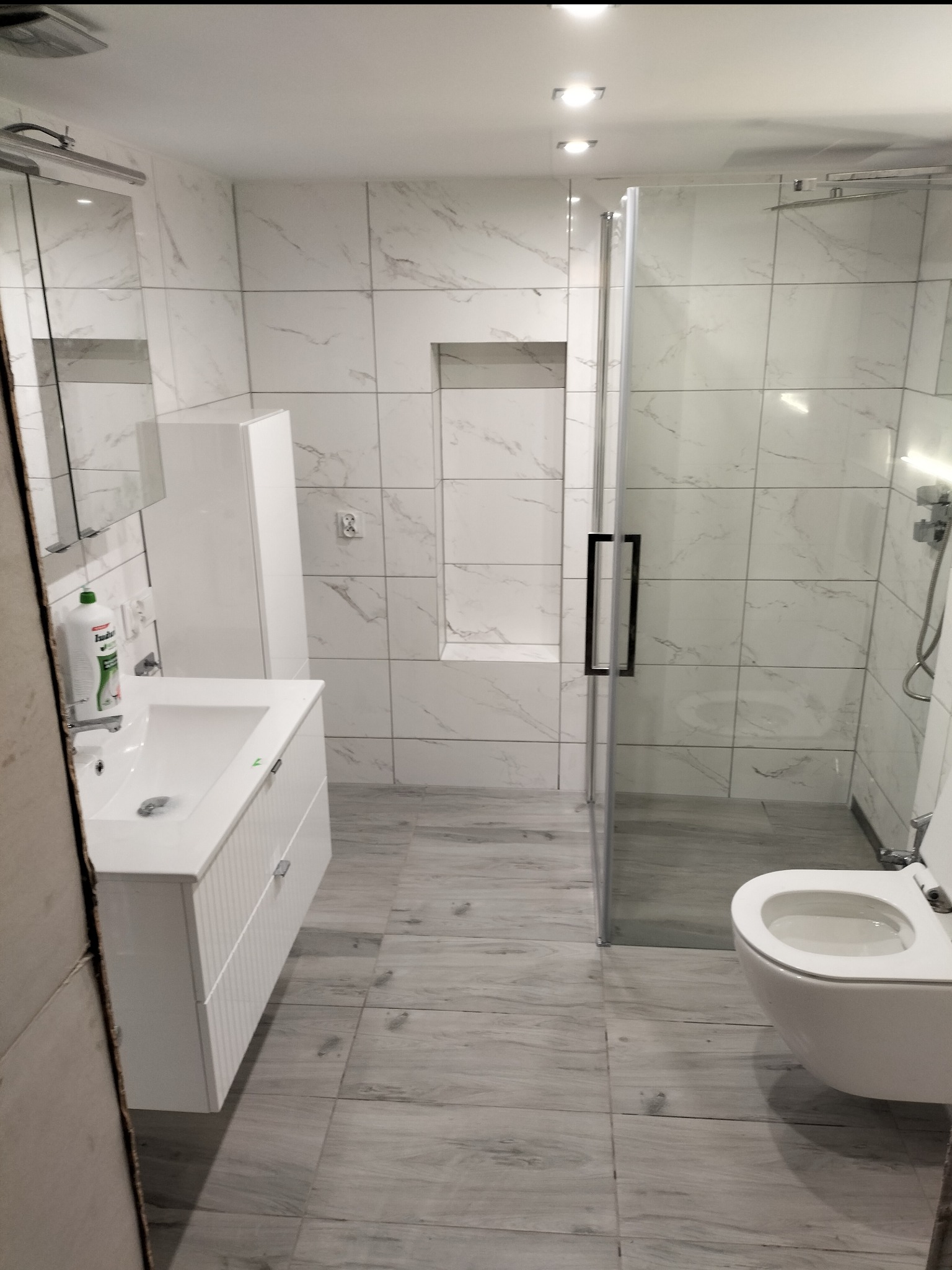 Nowoczesna łazienka z białymi marmurkowymi płytkami, kabiną prysznicową ze szklanymi drzwiami z czarnym uchwytem, białą umywalką z szafką i wiszącą toaletą, oświetlona punktowymi lampami sufitowymi.