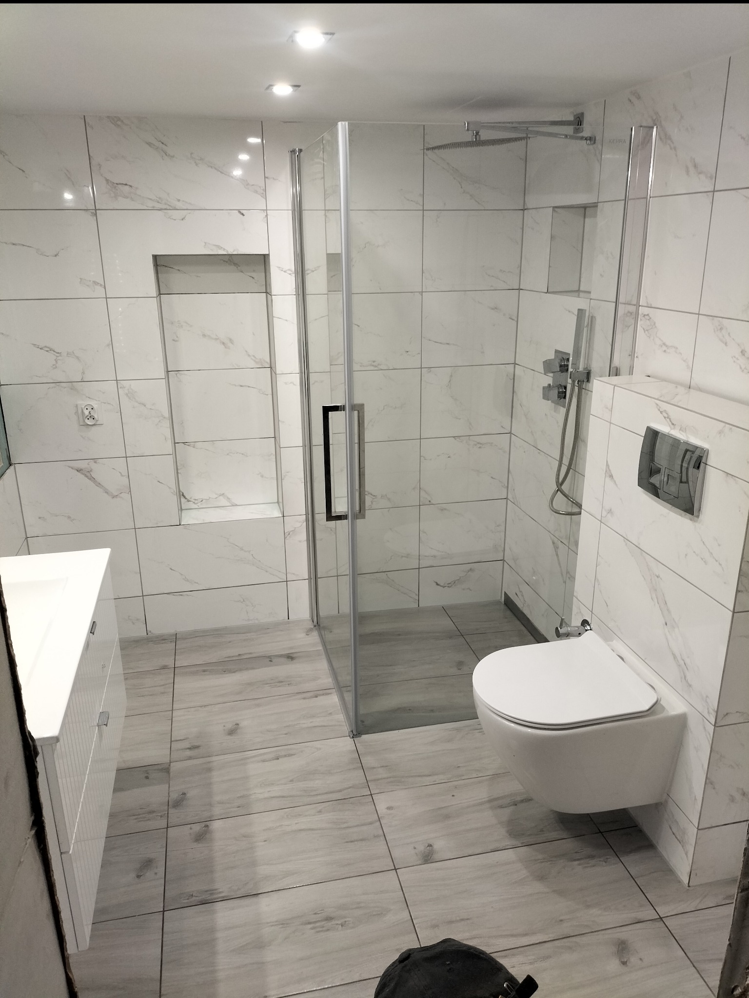 Nowoczesna łazienka z marmurowymi płytkami, kabiną prysznicową ze szklanymi drzwiami, wiszącą toaletą i umywalką z szafką. Widoczne chromowane baterie i deszczownica. Na podłodze płytki imitujące...