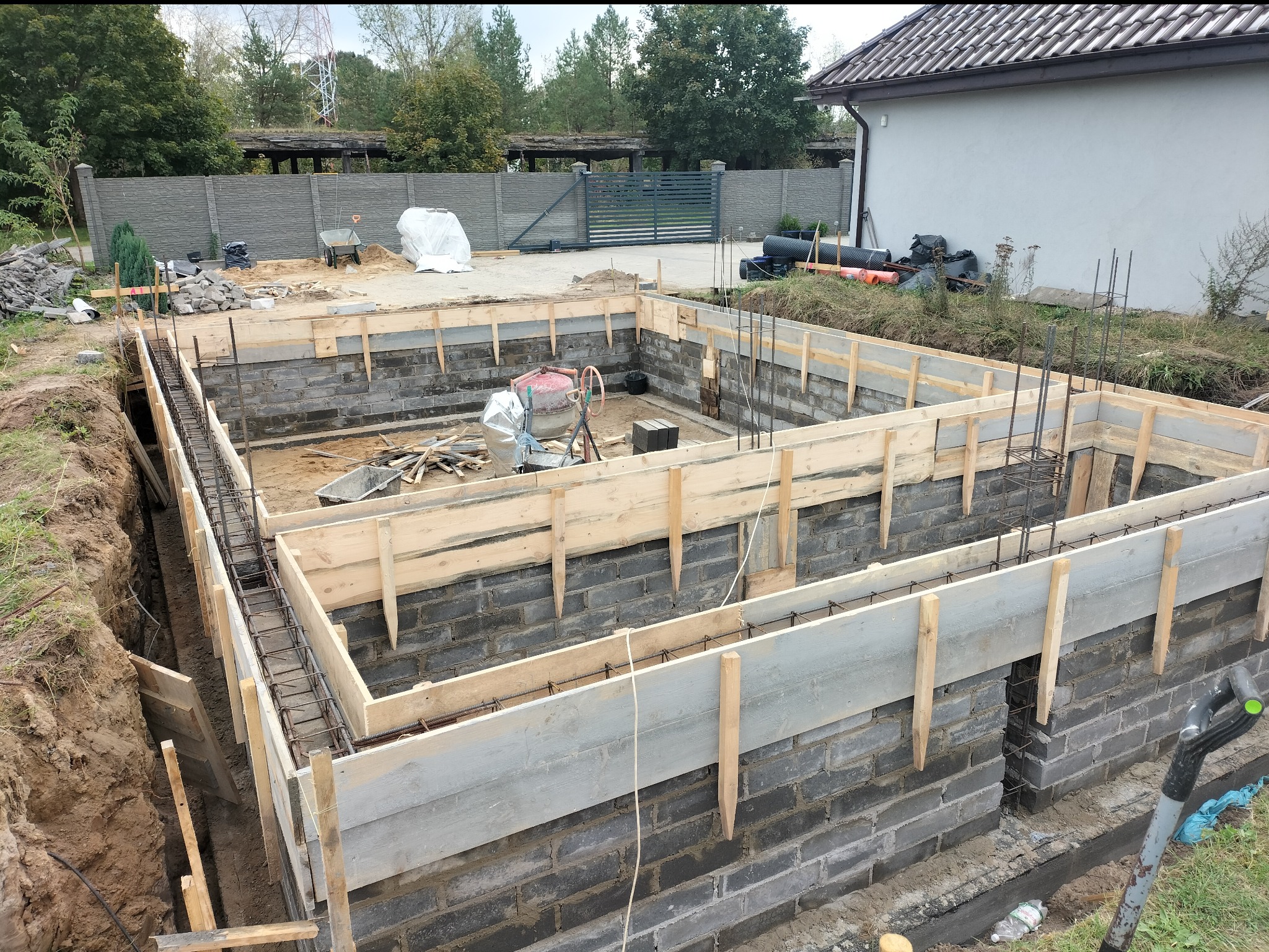 Budowa fundamentów domu jednorodzinnego: szalunek z desek, zbrojenie stalowe, bloczki betonowe, mieszalnik do betonu w tle, widok z góry.