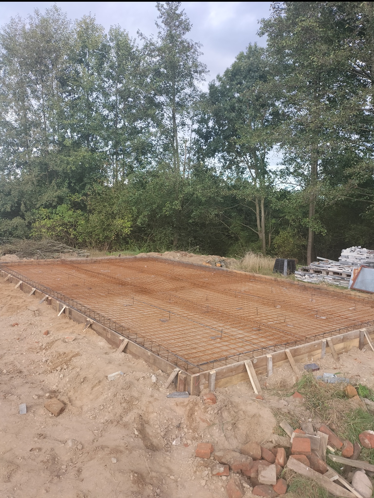 Przygotowanie zbrojenia i szalunku pod płytę fundamentową na gruncie piaszczystym, widoczne drzewa w tle.