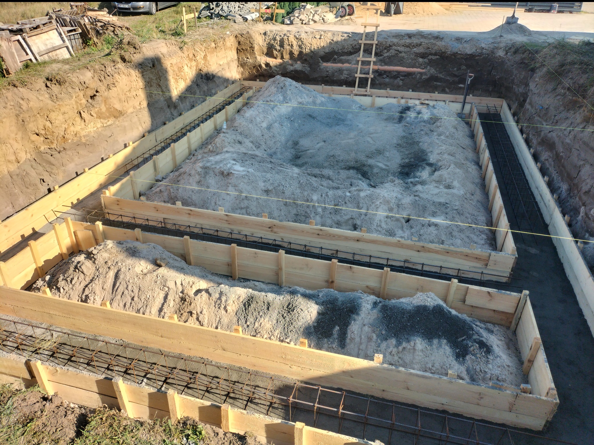Wykop pod fundamenty domu z drewnianym szalunkiem i zbrojeniem, wypełniony częściowo piaskiem i żwirem, widok z góry.