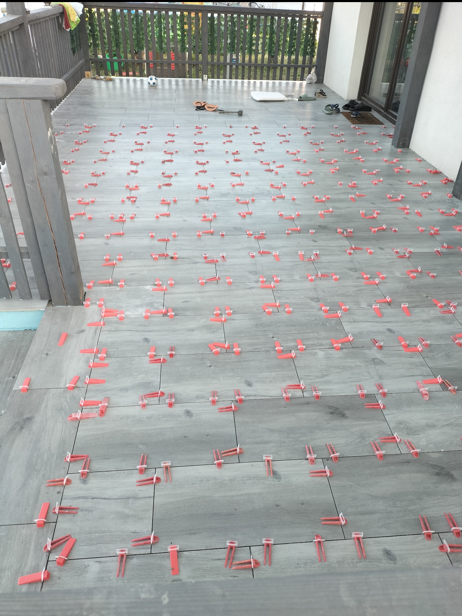 Układanie szarych płytek imitujących drewno na tarasie z użyciem czerwonych klipsów poziomujących, widok z góry na fragment tarasu z piłką i butami w tle.