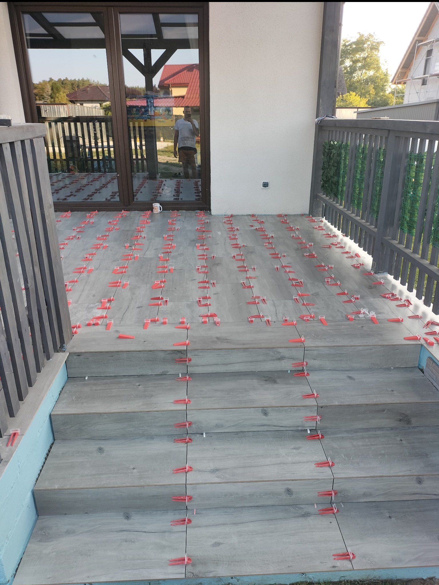 Układanie szarych płytek imitujących drewno na schodach i tarasie z użyciem czerwonych klinów poziomujących, widoczny kubek na tarasie i częściowo odbicie osoby w szklanych drzwiach.