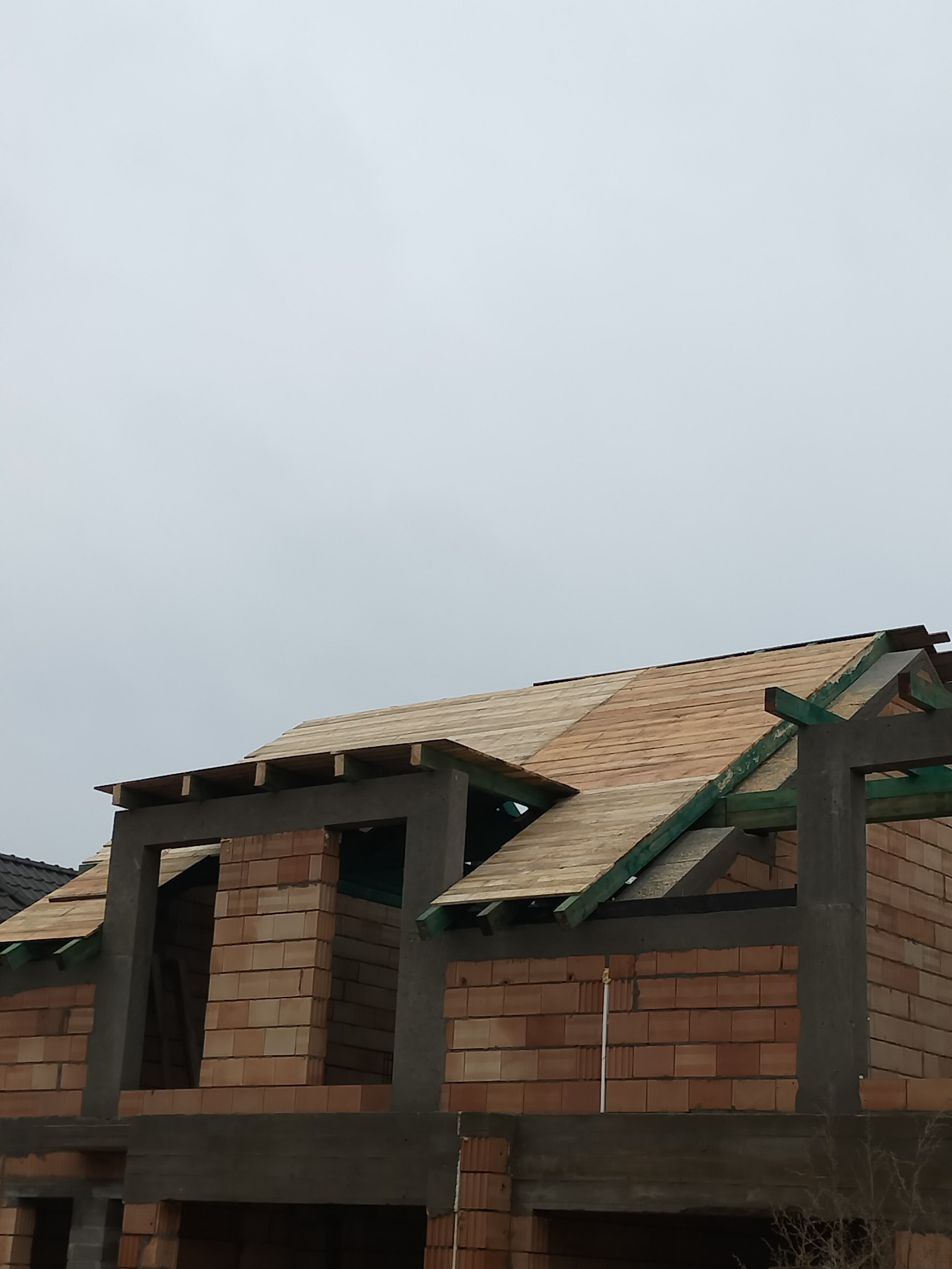 Surowa konstrukcja dachu z deskowaniem na ceglanym domu w trakcie budowy, widoczne elementy betonowe i zielone krokwie, pochmurne niebo w tle.