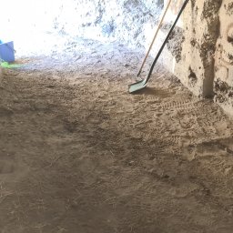 SOLID WORK Grzegorz Nieleszczuk - Wnętrze betonowego bunkra po wstępnym uprzątnięciu, widoczne ślady butów na ziemi, oparta o ścianę łopata i grabie, w tle niebieskie wiadro.