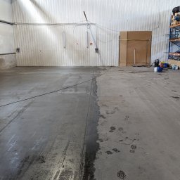 SOLID WORK Grzegorz Nieleszczuk - Częściowo wyczyszczona betonowa posadzka w hali magazynowej, widoczne ślady czyszczenia i ślady butów, w tle regały z towarem.