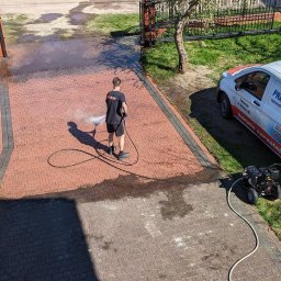SOLID WORK Grzegorz Nieleszczuk - Mężczyzna w czarnym ubraniu czyści czerwoną kostkę brukową za pomocą myjki ciśnieniowej; widoczny efekt czyszczenia, w tle brama i zaparkowany bus firmy sprzątającej.