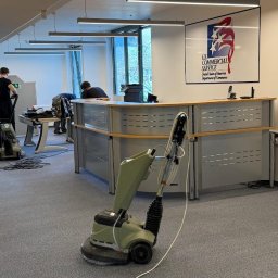 SOLID WORK Grzegorz Nieleszczuk - Pranie wykładziny w biurze US Commercial Service. Widoczne maszyny czyszczące i pracownicy w trakcie pracy na szarej wykładzinie biurowej.