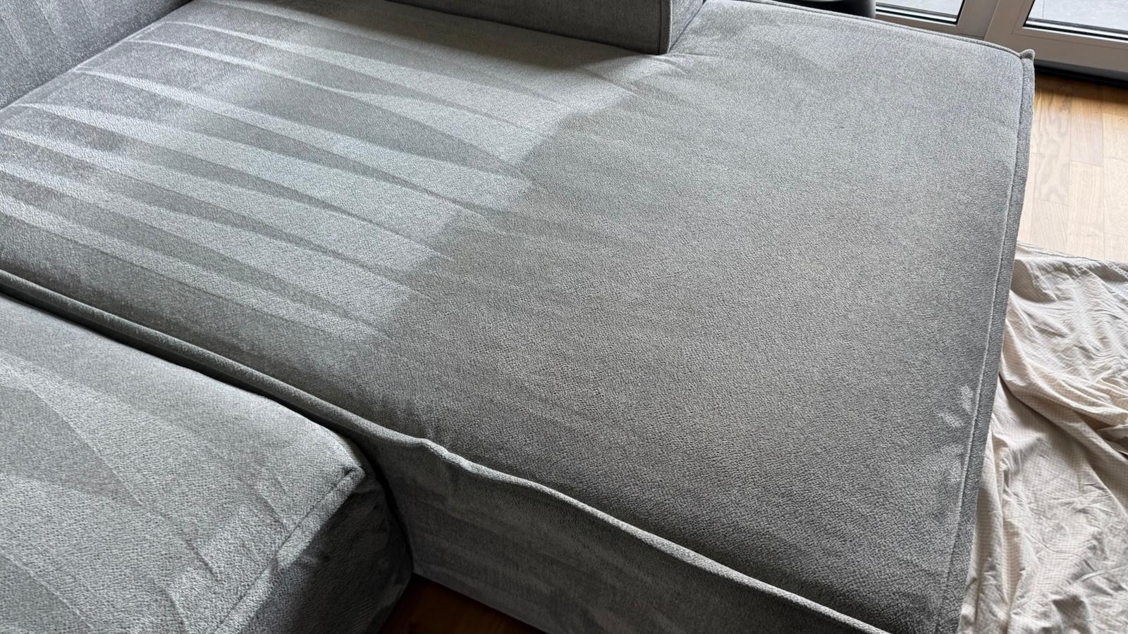 Fragment szarej sofy z widocznymi śladami prania tapicerki, ukazujący różnicę w czystości między wypraną a niewypraną częścią. Widoczna faktura materiału i fragment podłogi.