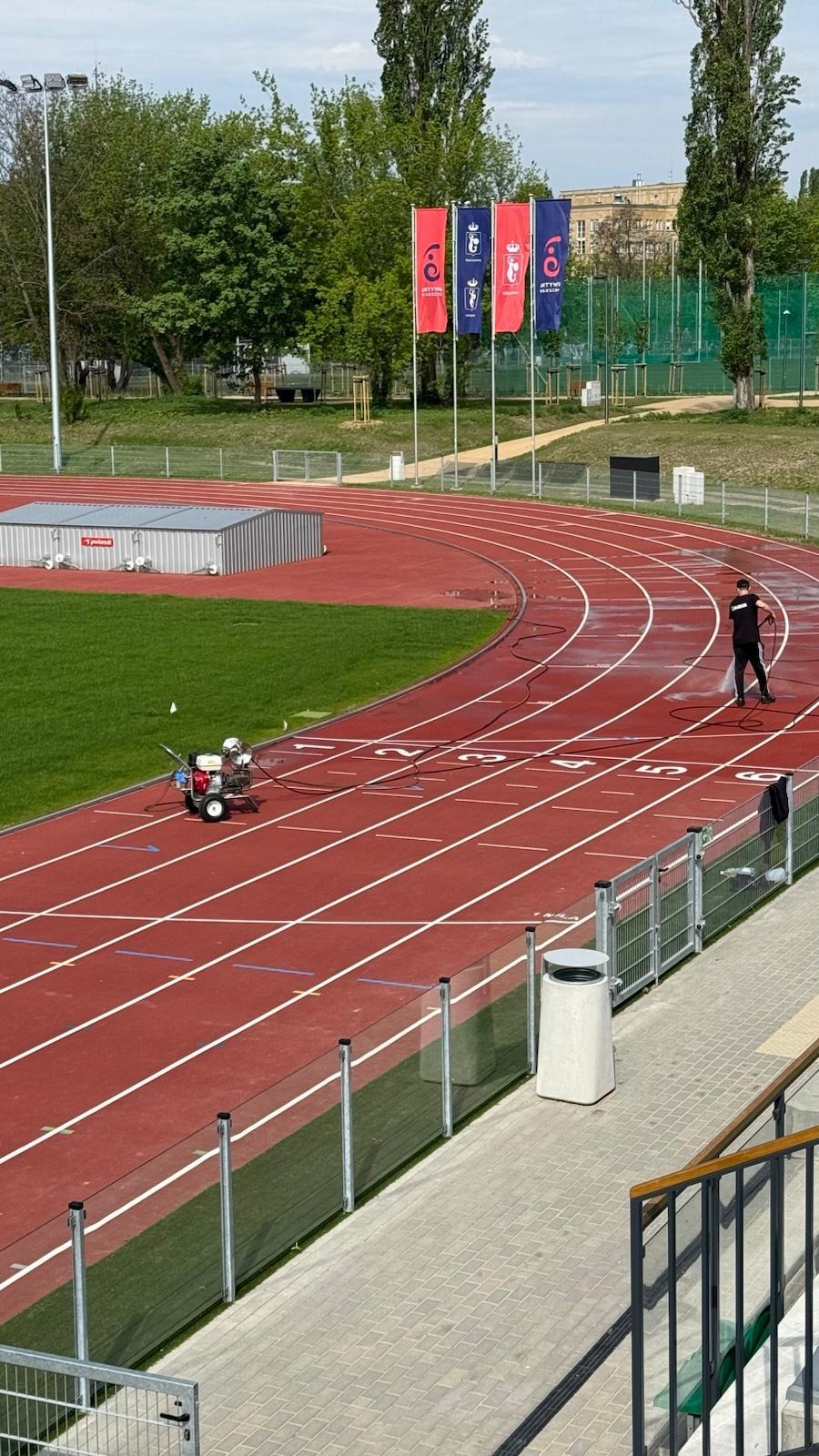 Czyszczenie czerwonej bieżni na stadionie sportowym za pomocą myjki ciśnieniowej. Pracownik w czarnym ubraniu w trakcie pracy. Widoczne flagi i drzewa w tle.