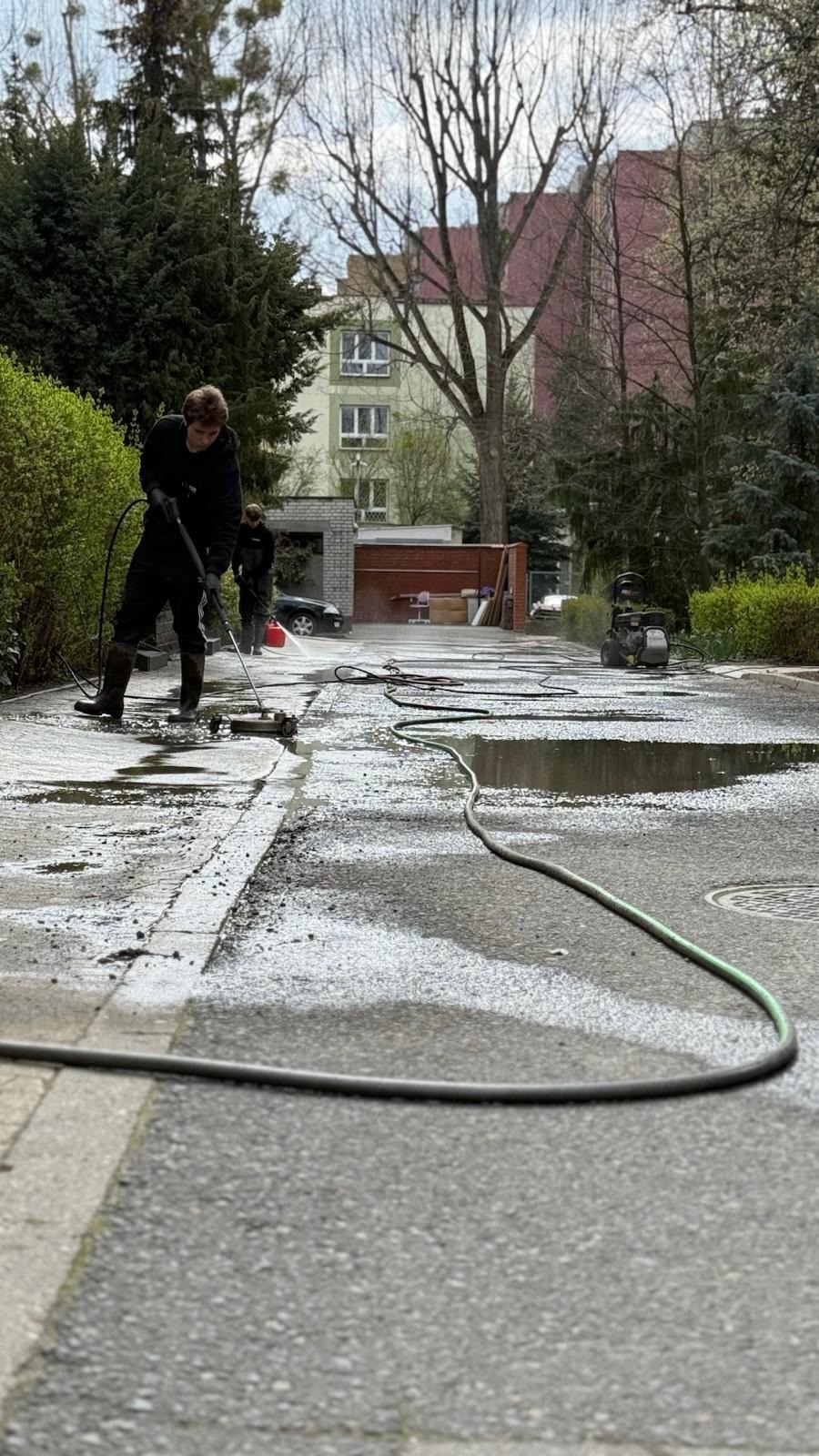 Dwóch mężczyzn czyści kostkę brukową za pomocą myjek ciśnieniowych. W tle budynek mieszkalny i drzewa. Woda i piana na czyszczonej powierzchni.