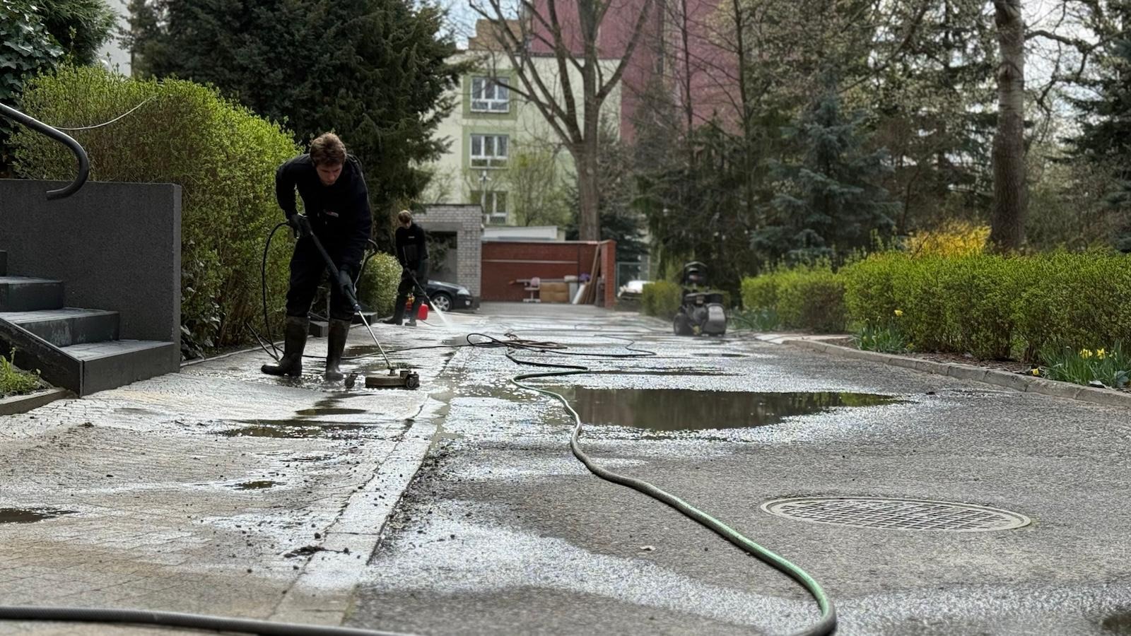 Dwaj mężczyźni w czarnych strojach czyszczą kostkę brukową za pomocą myjek ciśnieniowych. Widoczne kałuże i wąż ogrodowy. Tło stanowią budynki i zieleń.
