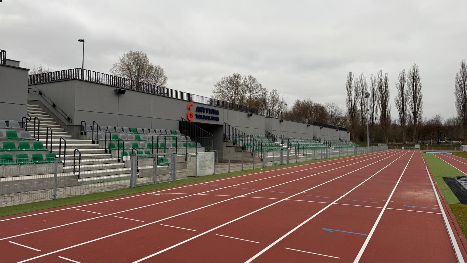 Czerwona bieżnia lekkoatletyczna z białymi liniami, trybuny z zielonymi krzesełkami, szare budynki w tle i pochmurne niebo. Widok z perspektywy bieżni.