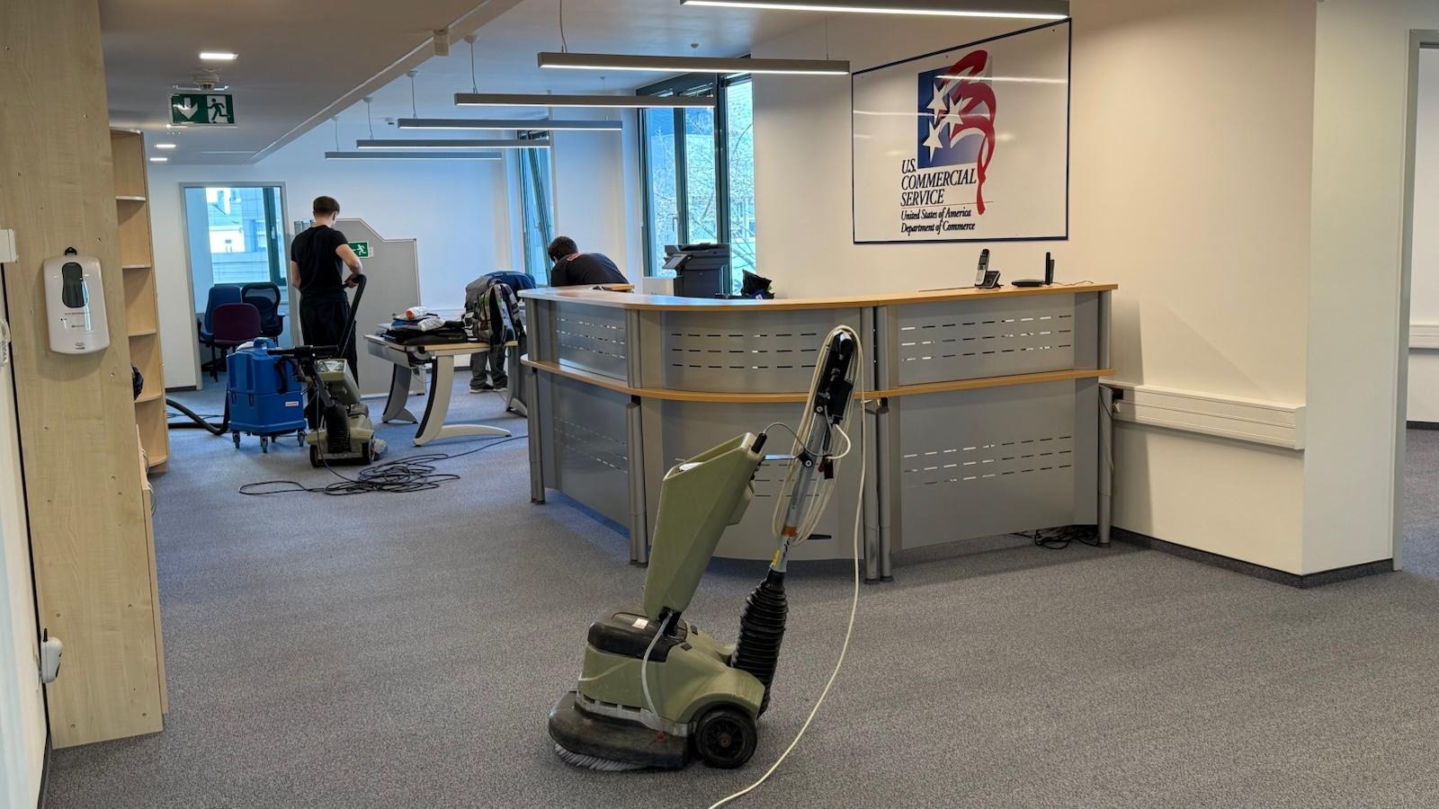 Pranie wykładziny w biurze US Commercial Service. Widoczne maszyny czyszczące i pracownicy w trakcie pracy na szarej wykładzinie biurowej.