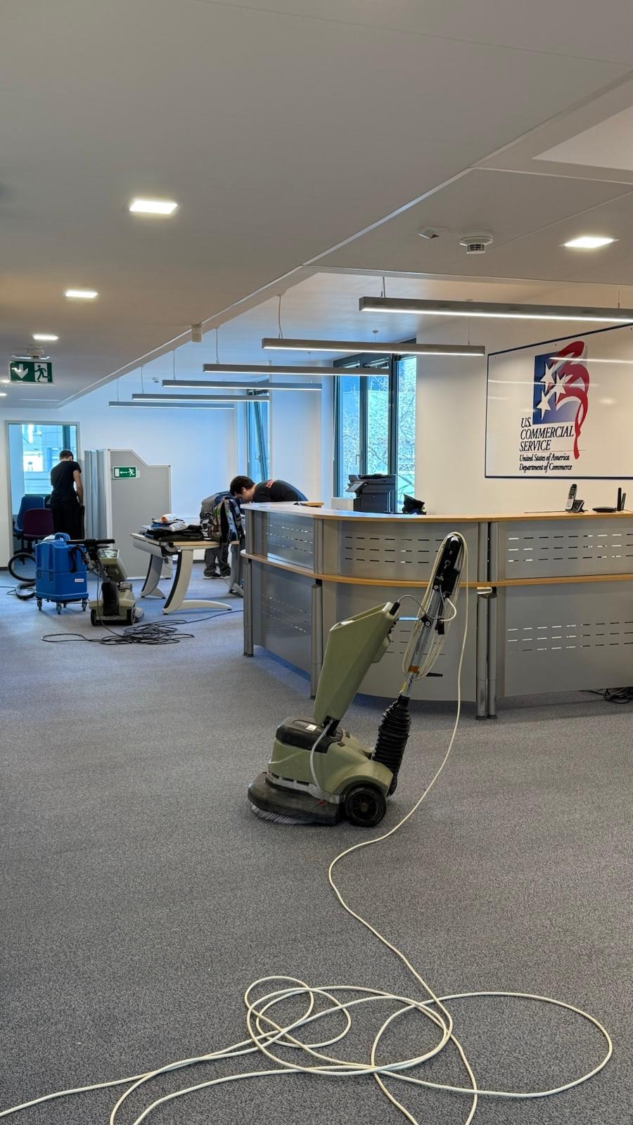 Pranie wykładziny w biurze: maszyna czyszcząca na szarym dywanie, widoczna recepcja z logo US Commercial Service i pracownicy w tle. Jasne oświetlenie.