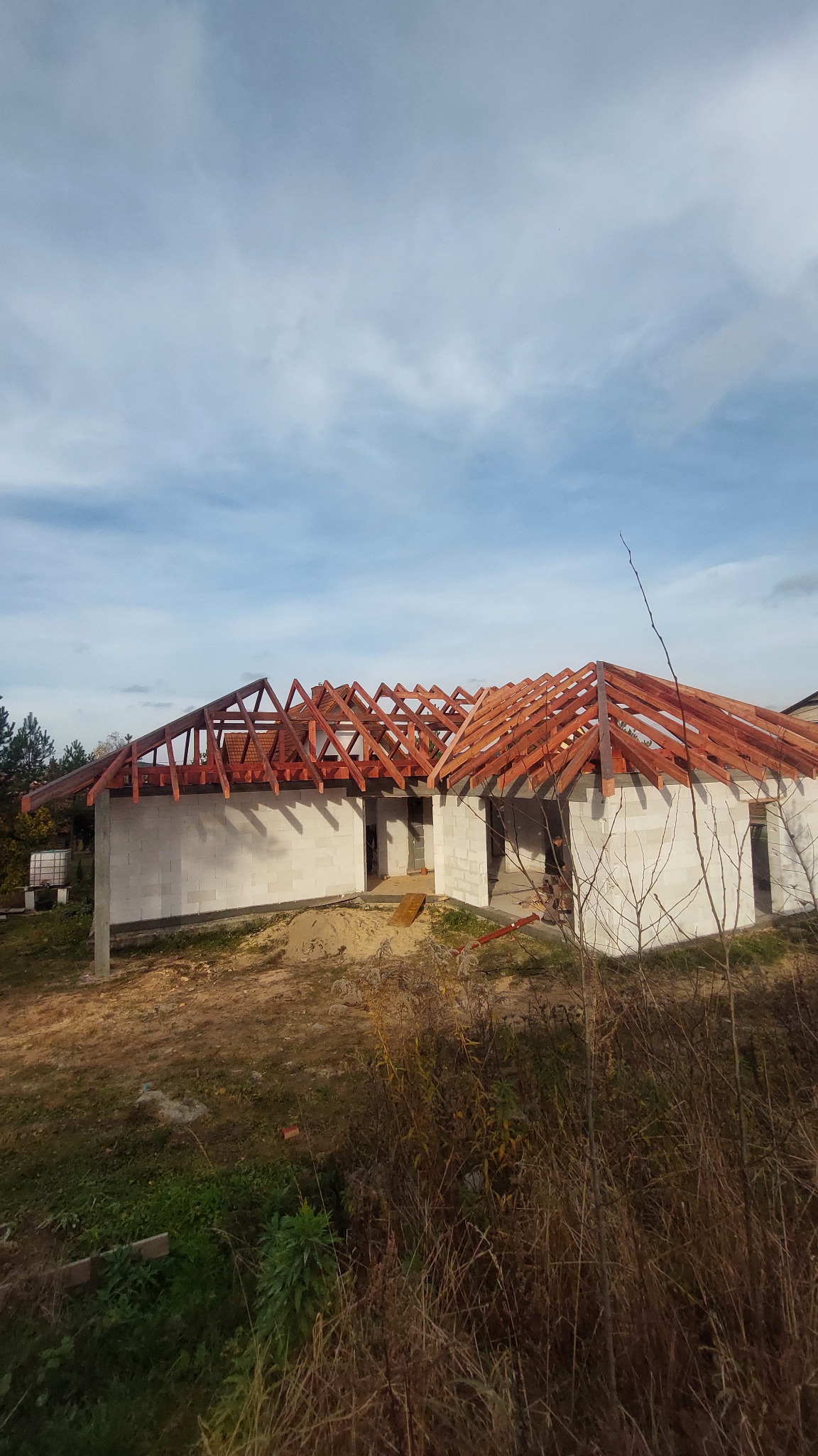 Surowy stan budowy domu jednorodzinnego z widoczną konstrukcją dachu z drewna, ściany z białych bloczków, teren budowy z roślinnością.