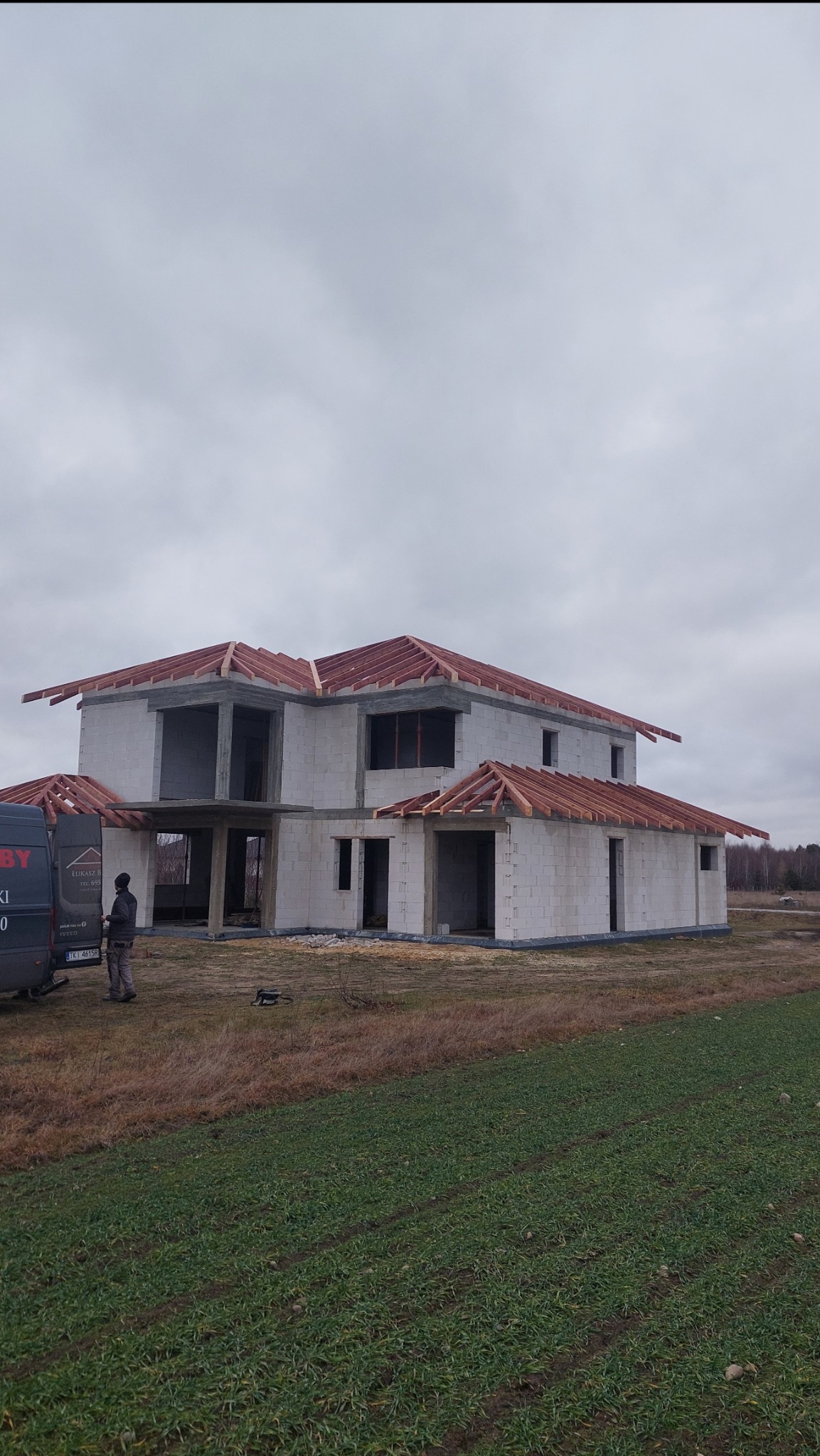 Budowa domu jednorodzinnego w stanie surowym otwartym, widoczna częściowo wykonana konstrukcja dachu z drewnianych krokwi, na tle pochmurnego nieba i zielonego pola, obok budynku stoi bus firmy...
