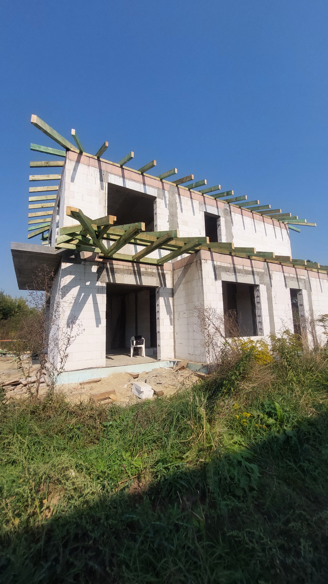 Surowy stan domu w budowie z widoczną konstrukcją drewnianą dachu, ściany z białych bloczków, otoczenie zarośnięte trawą i chwastami, krzesło wewnątrz budynku.