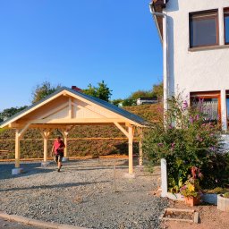 DANDREW SPÓŁKA Z OGRANICZONĄ ODPOWIEDZIALNOŚCIĄ - Drewniana wiata garażowa z szarym dachem na żwirowym podłożu, z mężczyzną wchodzącym do środka. W tle dom z białymi ścianami i zielenią.