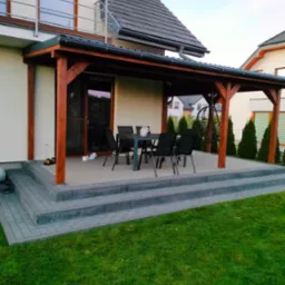 Zadaszony taras z drewnianą konstrukcją i schodami z kostki brukowej, wyposażony w stół i krzesła. W tle zielony trawnik i tuje.