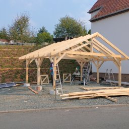 DANDREW SPÓŁKA Z OGRANICZONĄ ODPOWIEDZIALNOŚCIĄ - Konstrukcja drewniana z jasnego drewna w trakcie budowy, widoczne narzędzia i drabiny, dom w tle, słoneczny dzień.