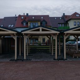 DANDREW SPÓŁKA Z OGRANICZONĄ ODPOWIEDZIALNOŚCIĄ - Trzy drewniane, połączone wiaty garażowe z czarnymi dachami na posesji z kostką ażurową, na tle budynków mieszkalnych w pochmurny dzień.