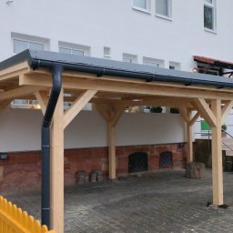 DANDREW SPÓŁKA Z OGRANICZONĄ ODPOWIEDZIALNOŚCIĄ - Nowoczesna, drewniana konstrukcja z szarą blachą trapezową, prawdopodobnie zadaszenie lub wiata, przylegająca do budynku z oknami. Widoczny system rynnowy.