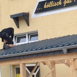 DANDREW SPÓŁKA Z OGRANICZONĄ ODPOWIEDZIALNOŚCIĄ - Dekarz montuje dach z blachy trapezowej na drewnianej konstrukcji. Widoczne narzędzia dekarskie i fragment budynku z oknami. Ujęcie z dołu, z bliska.