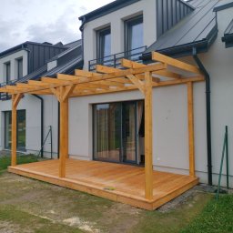 DANDREW SPÓŁKA Z OGRANICZONĄ ODPOWIEDZIALNOŚCIĄ - Drewniana pergola z tarasem przy nowoczesnym domu z grafitowym dachem. Jasne drewno konstrukcji kontrastuje z bielą elewacji. Widoczne złączenia ciesielskie.