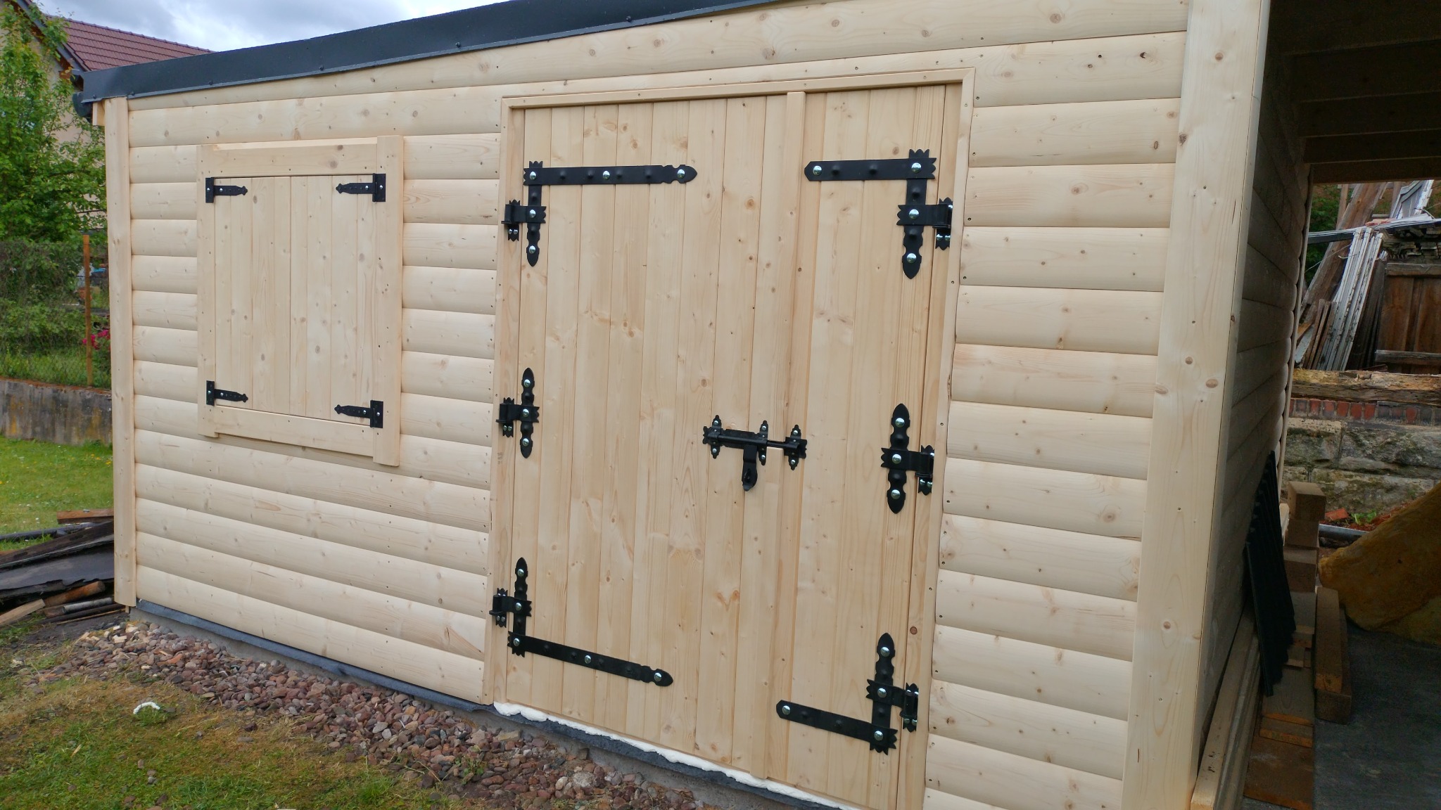 Drewniany domek narzędziowy z zamkniętymi okiennicami i ozdobnymi, kutymi zawiasami. Ściany z jasnych bali, czarny dach. Widok z zewnątrz, z boku.