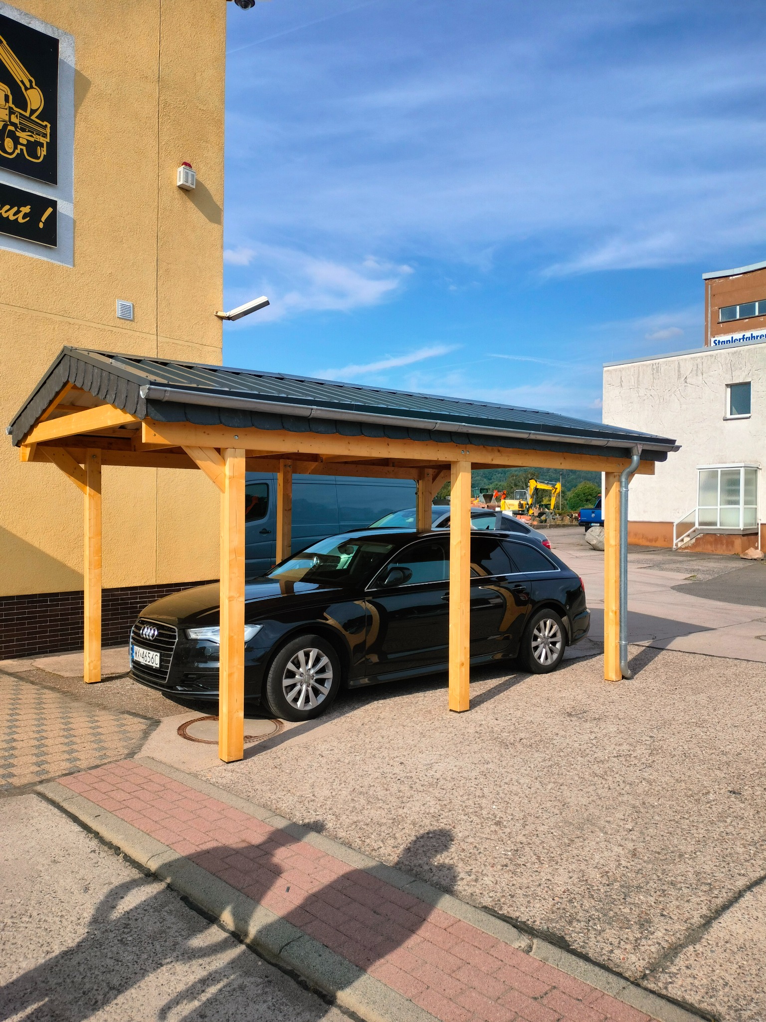 Czarny samochód osobowy pod zadaszeniem z drewna i ciemnej dachówki, zaparkowany na betonowym placu. W tle budynek z żółtą elewacją i niebieskie niebo.