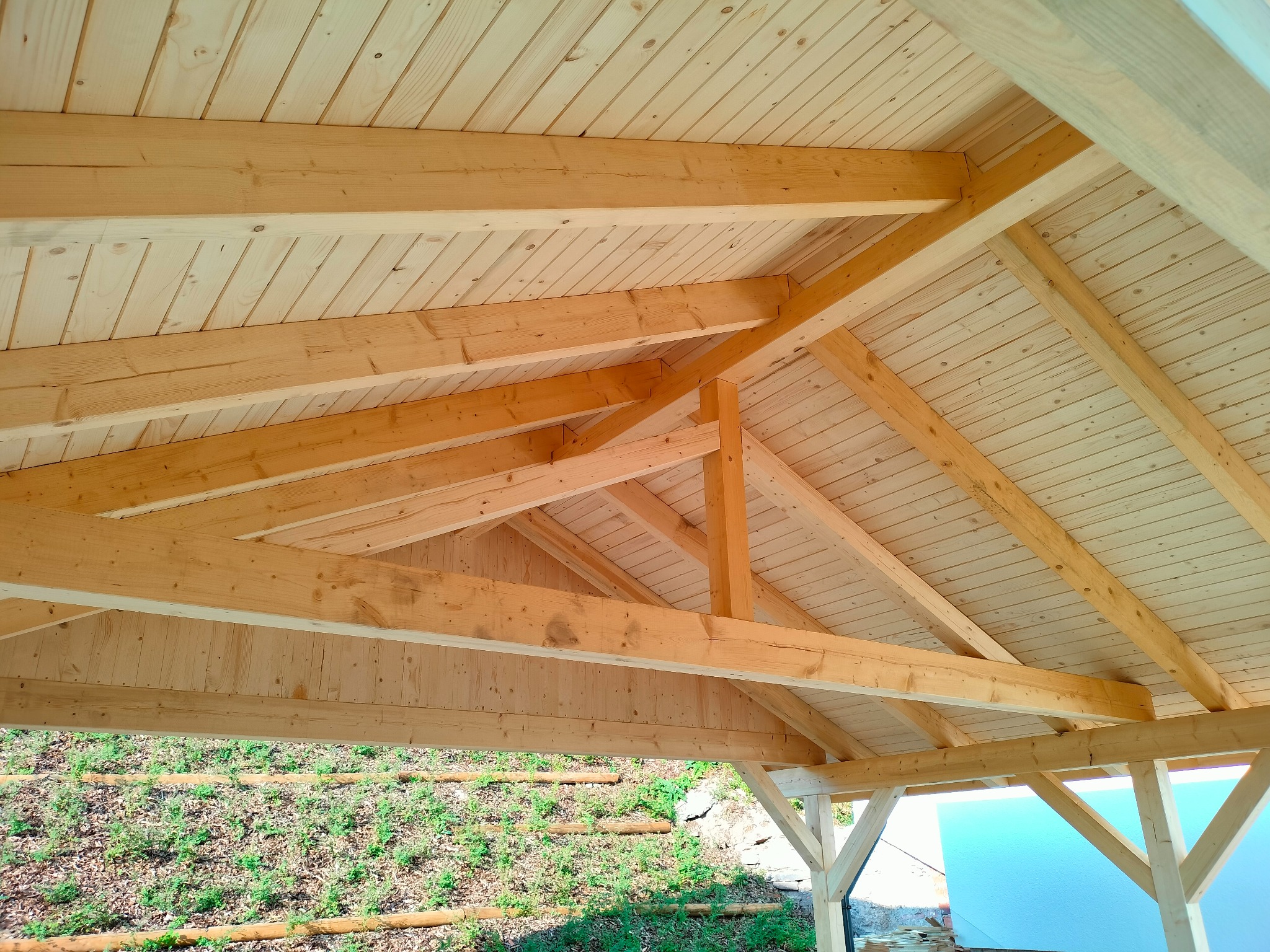 Jasna więźba dachowa z drewna świerkowego, widok od dołu, z fragmentem zielonego zbocza w tle. Element konstrukcyjny z widocznymi belkami i deskowaniem.