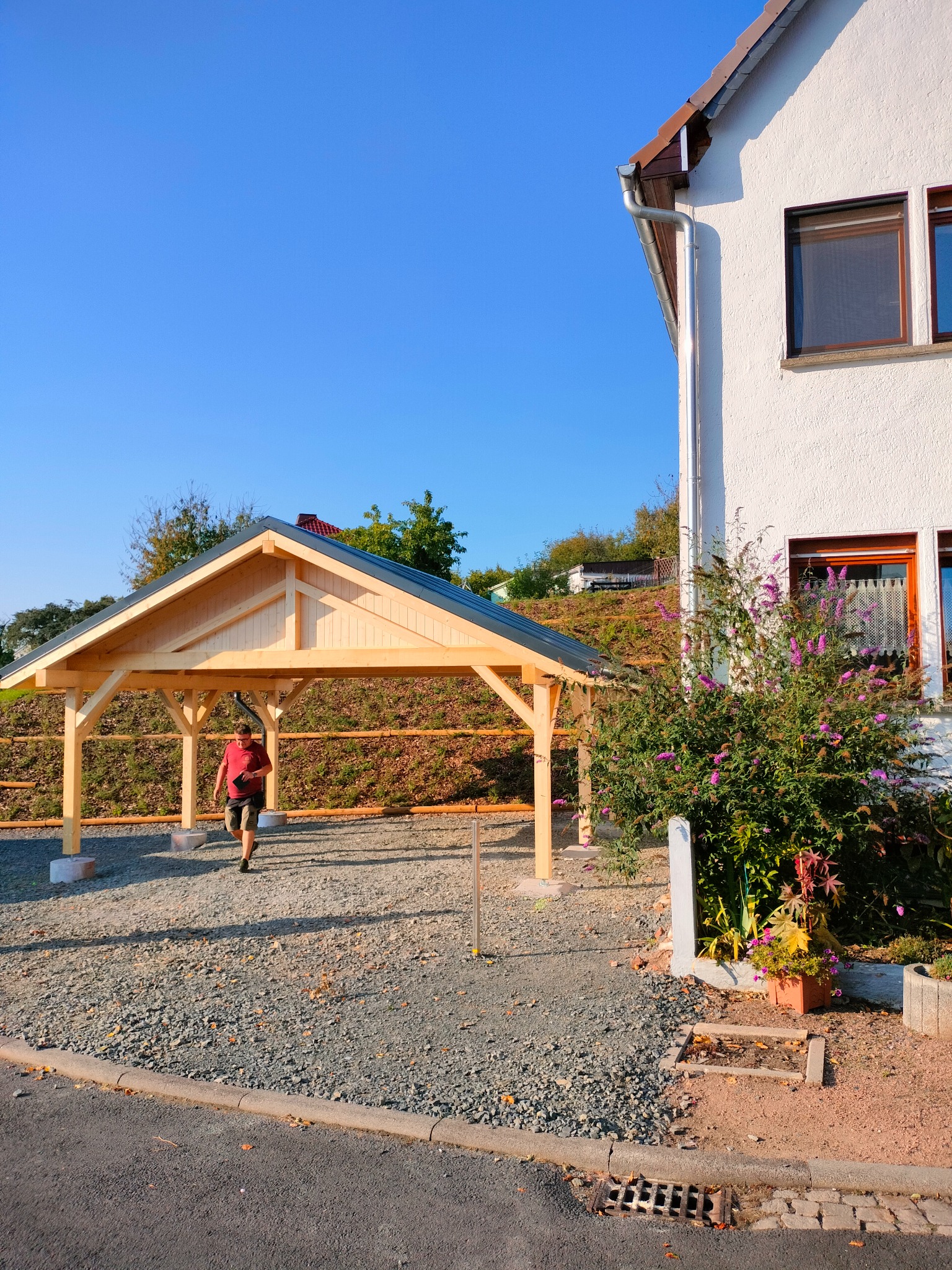 Drewniana wiata garażowa z szarym dachem na żwirowym podłożu, z mężczyzną wchodzącym do środka. W tle dom z białymi ścianami i zielenią.